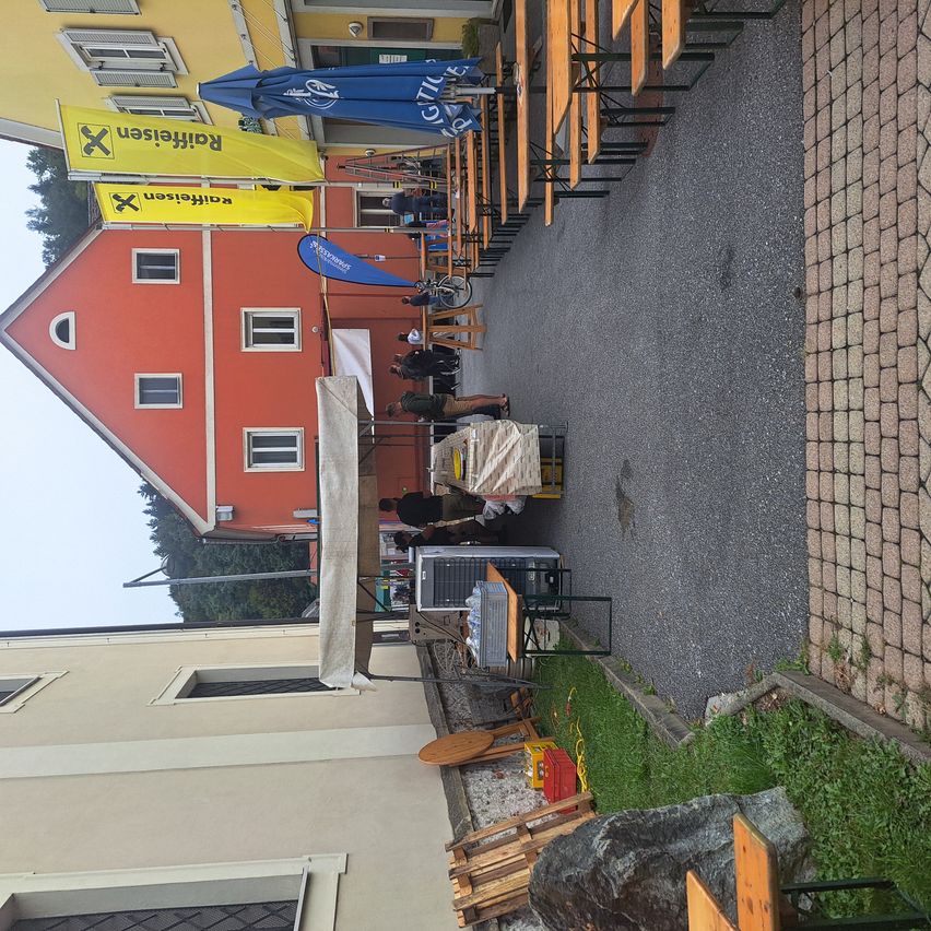 Ein Außenbereich mit mehreren Holzbänken und Sonnenschirmen. Zwei Personen richten einen Tisch mit einer Klimaanlage ein. Ein Gebäude mit mehreren Fenstern und gelben Bannern befindet sich in der Nähe.