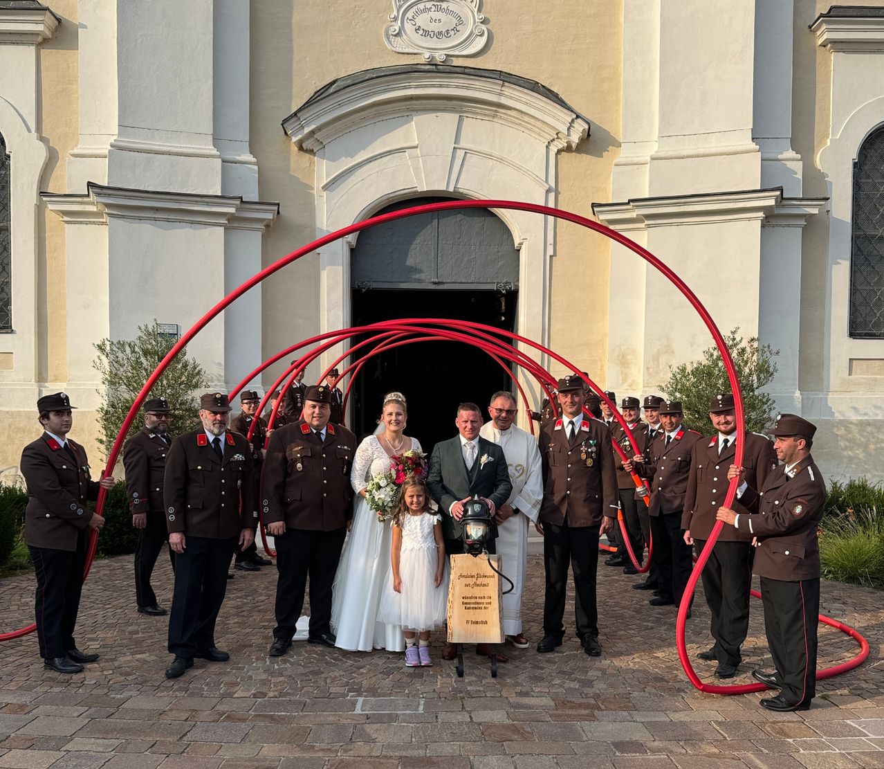 Eine Hochzeitszeremonie findet vor einer Kirche mit uniformierten Personen statt. Die Braut und der Bräutigam stehen mit einem kleinen Mädchen, und ein Mann hält einen Feuerlöscher.