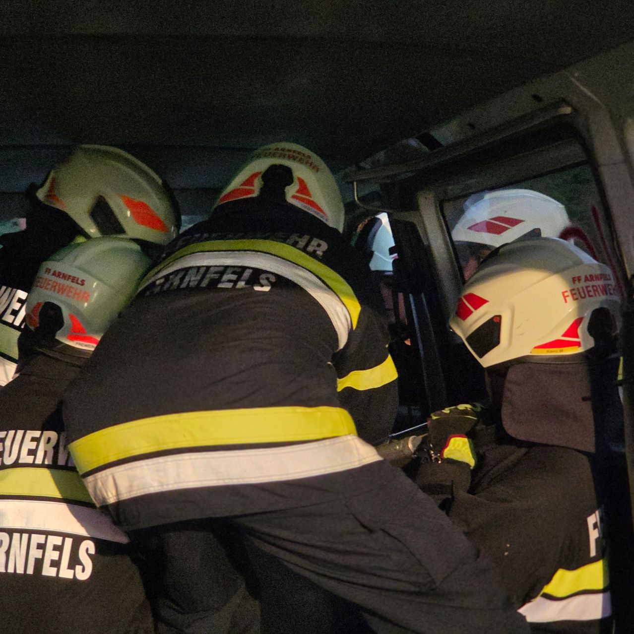 Ein Team von Feuerwehrleuten in voller Ausrüstung, einschließlich Helmen mit der Aufschrift 'Feuerwehr', rettet jemanden aus einem Fahrzeug, wobei ein Feuerwehrmann ein Werkzeug hält.