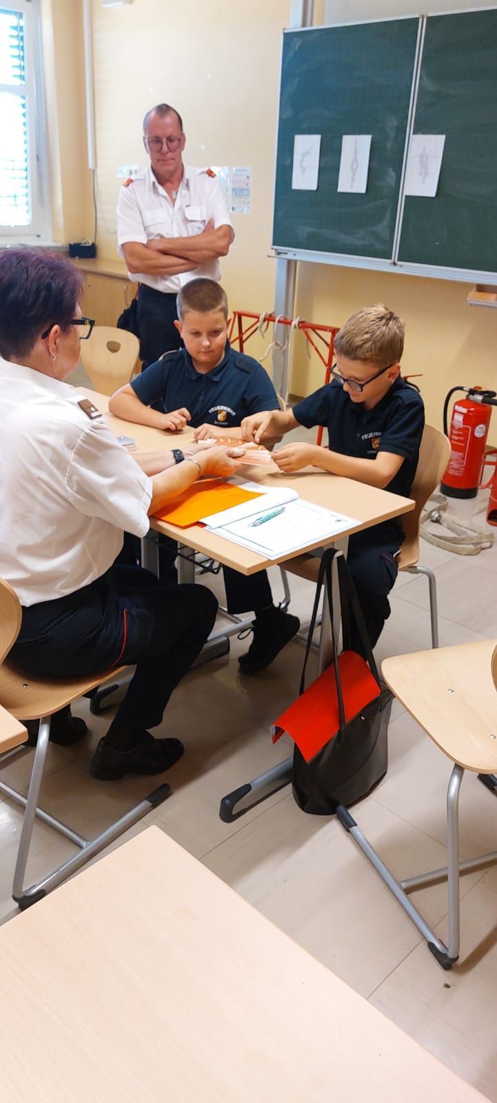 Eine Frau und zwei Jungen in Uniformen sitzen an einem Tisch, ein Junge hält eine Karte. Papiere und ein Stift liegen auf dem Tisch. Ein Feuerlöscher steht hinter ihnen.