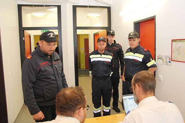 Fünf Männer in Uniform, einer sitzt und vier stehen, befinden sich in einer Besprechung. Einer der stehenden Männer schaut sich einen Laptop auf dem Tisch an.
