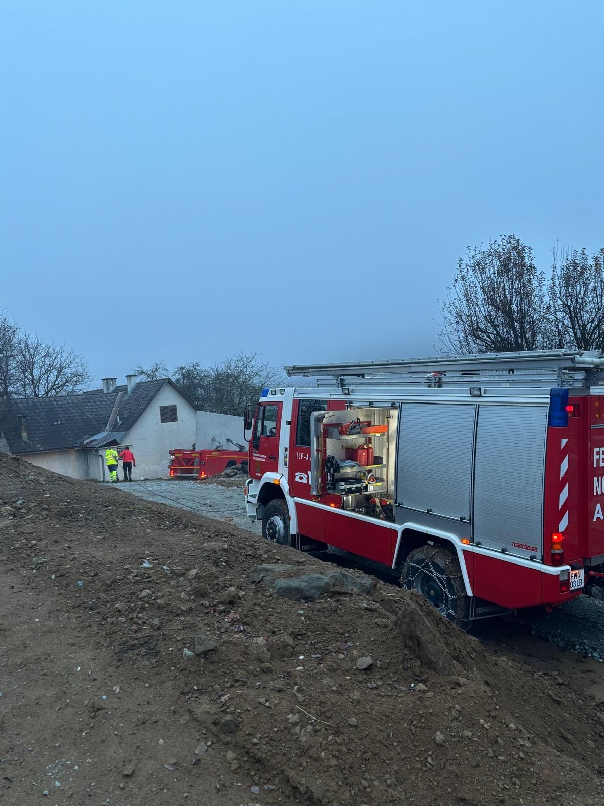 Ein roter Feuerwehrwagen steht auf einer Erdstraße in der Nähe eines Hauses. Zwei Feuerwehrleute stehen in der Nähe, und ein weiterer LKW ist hinter dem Haus geparkt.