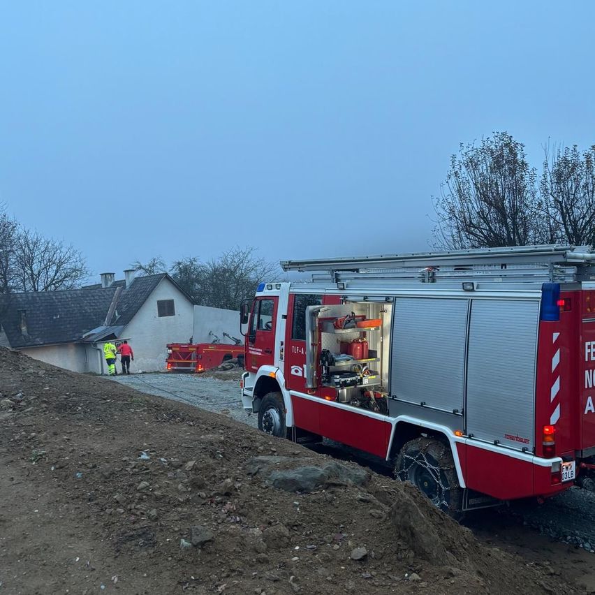 Ein roter Feuerwehrwagen steht auf einer Erdstraße in der Nähe eines Hauses. Zwei Feuerwehrleute stehen in der Nähe, und ein weiterer LKW ist hinter dem Haus geparkt.