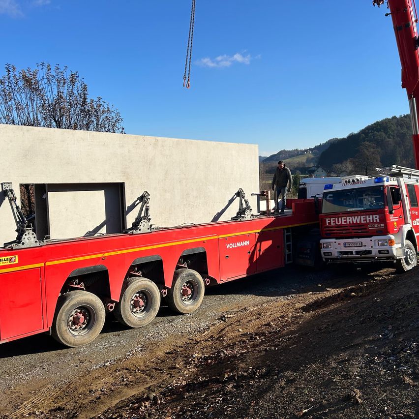 Ein großer roter LKW mit einem Kran steht auf einer Straße und transportiert eine große Betonstruktur. Ein Mann steht auf dem LKW.