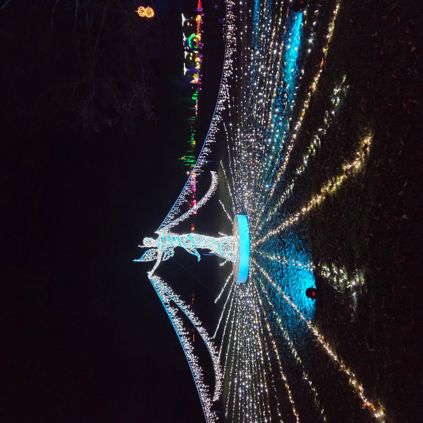 Eine große Engelsstatue mit Flügeln, geschmückt mit weißen und blauen Lichtern, ist nachts beleuchtet. Sie ist von einem Kreis blauer Lichter und zusätzlichen Lichtern im Hintergrund umgeben.