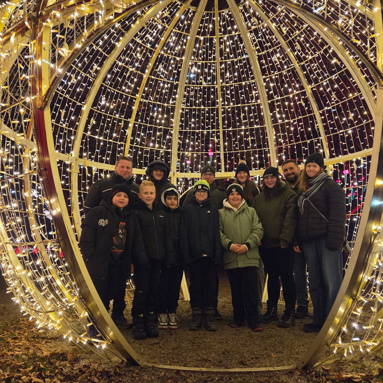 Eine Gruppe von Menschen, sowohl Erwachsene als auch Kinder, posiert für ein Foto in einer dekorativen, beleuchteten Kugel, umgeben von festlichen Lichtern.
