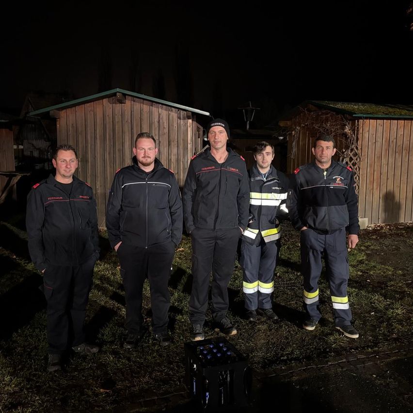 Fünf Feuerwehrleute in Uniform stehen bei Nacht vor Holzhütten, einer von ihnen hält einen schwarzen Kasten vor sich.