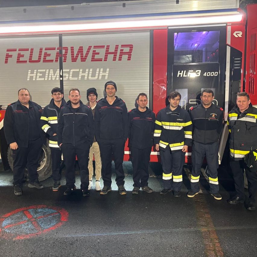 Eine Gruppe Feuerwehrleute in Uniform posiert vor einem Feuerwehrauto mit der Aufschrift 'Feuerwehr Heimschuh' und 'HLF3 4000'. Sie stehen in einer Reihe mit einem kreisförmigen Zeichen auf dem Boden.