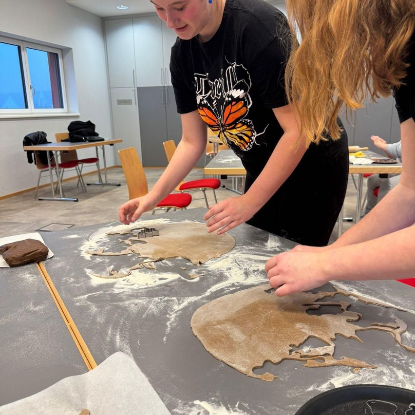 Zwei Frauen backen in einem Klassenzimmer. Eine Frau verwendet einen Ausstecher, um eine Form aus dem Teig zu schneiden. Eine andere Frau legt ein gerolltes Teigstück auf den Tisch.