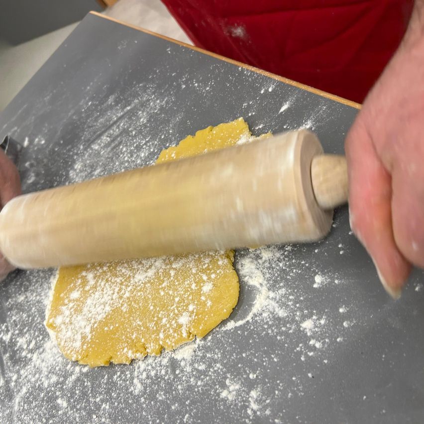 Eine Person rollt Teig auf einer grauen Oberfläche mit einem hölzernen Nudelholz aus. Der Teig ist mit Mehl bestreut.