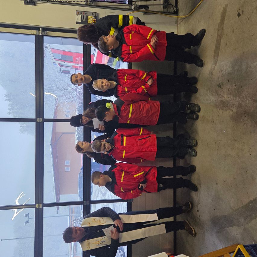 Eine Gruppe von Kindern in roten Jacken und schwarzen Hosen steht in einer Reihe, wahrscheinlich für ein Foto. Dahinter zeigt ein Glasfenster ein Gebäude und Bäume.