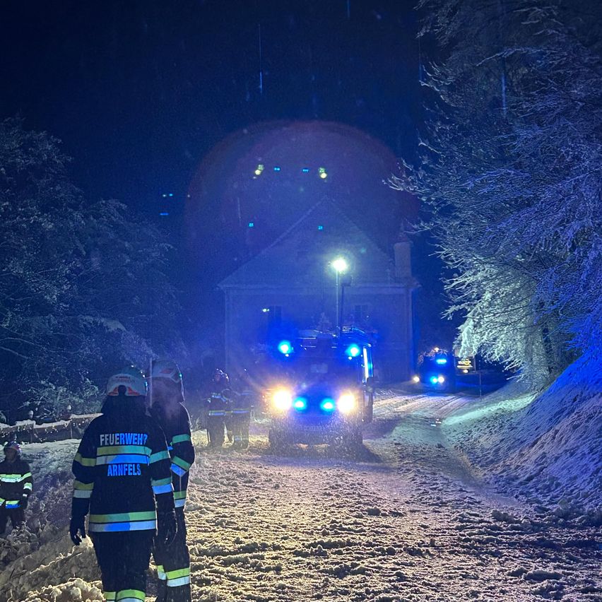Feuerwehrleute sind nachts auf einer verschneiten Straße, in der Nähe eines Gebäudes mit leuchtenden Lichtern. Zwei Fahrzeuge mit blauen Lichtern sind davor.