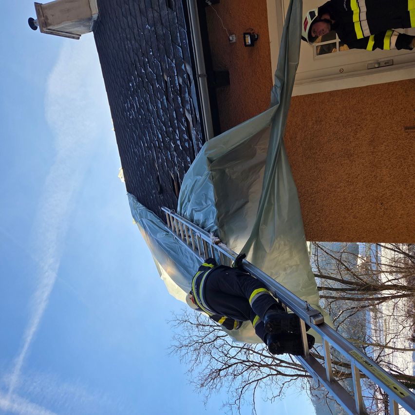 Zwei Feuerwehrleute in Uniform stehen auf einer Leiter, einer befestigt eine Plane am Dach. Das Gebäude hat eine braune Wand und eine weiße Tür.