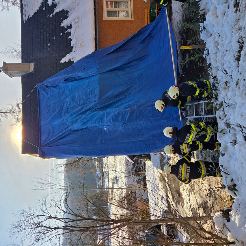 Eine Gruppe von Feuerwehrleuten in Uniform sichert eine blaue Plane über ein schneebedecktes Haus.