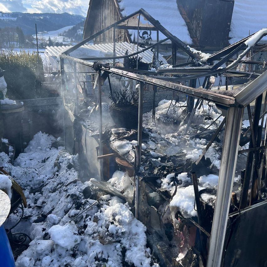 Ein Gewächshaus ist niedergebrannt und mit Schnee bedeckt. Rauch steigt aus den Überresten auf. Ein Haus ist im Hintergrund zu sehen.