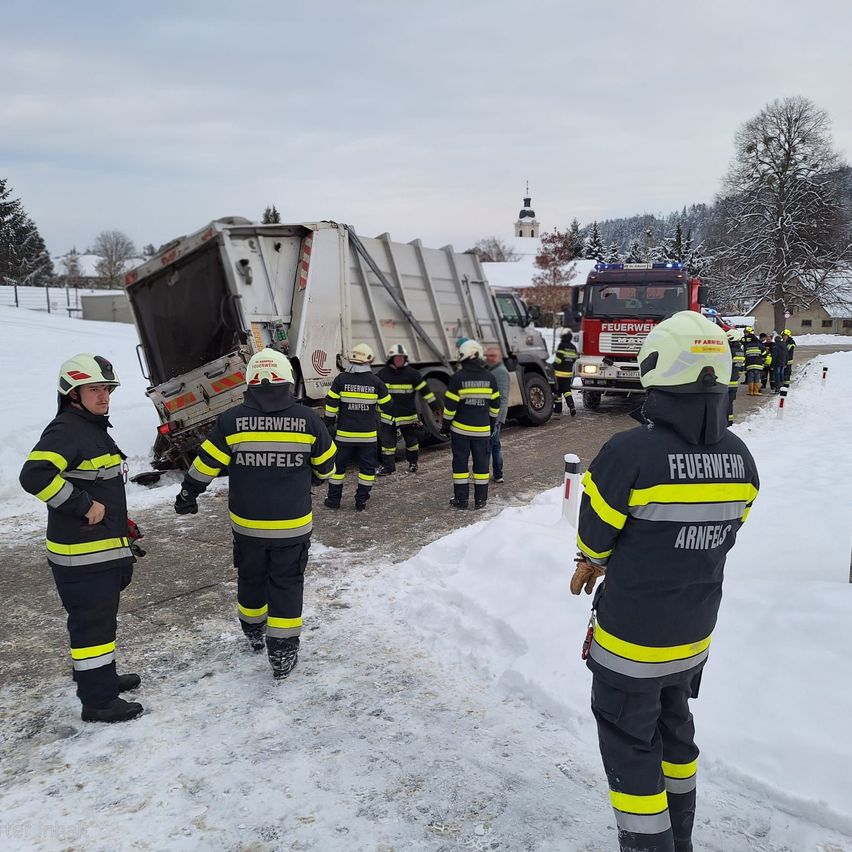 Feuerwehrleute in Uniformen stehen im Schnee neben einem umgestürzten Müllwagen. Ein Feuerwehrwagen ist in der Nähe geparkt.