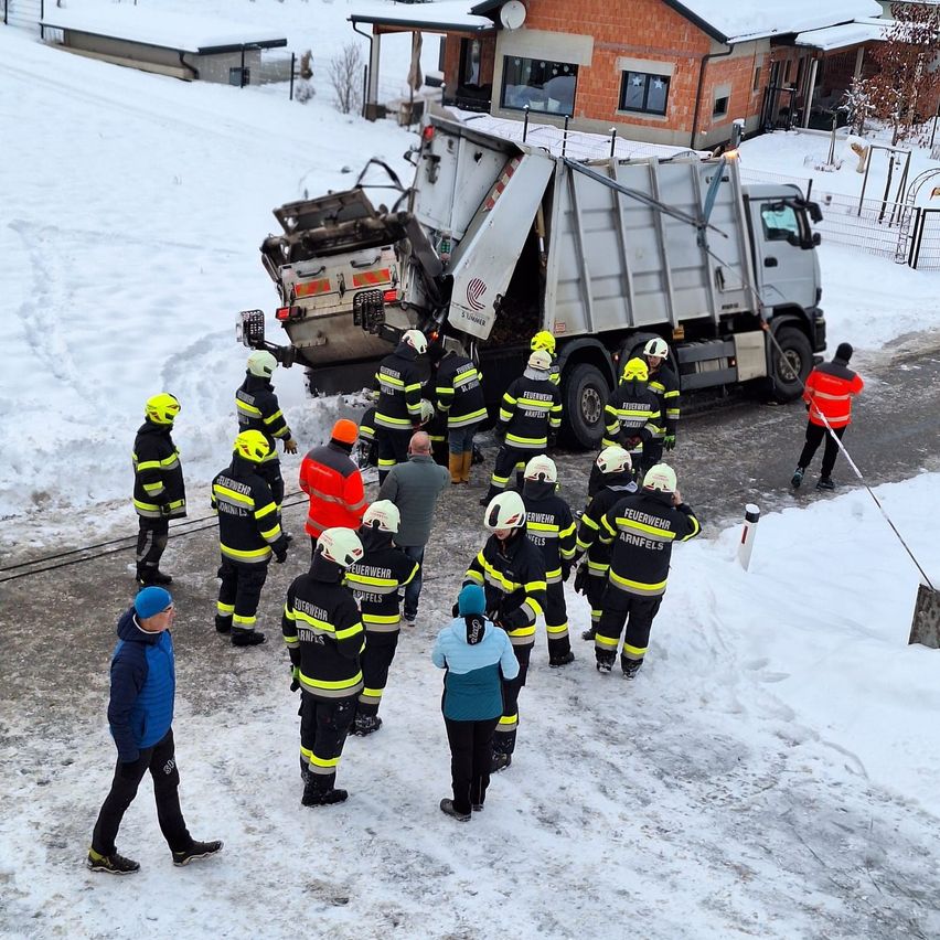 Feuerwehrleute in Schutzkleidung stehen auf einem verschneiten Gelände versammelt, mit einem beschädigten Lastwagen dahinter. Eine Person hält eine Schaufel.