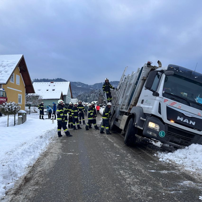 Eine Gruppe Feuerwehrleute in Winterkleidung hilft einem feststeckenden Lastwagen auf einer verschneiten Straße. Im Hintergrund sind schneebedeckte Häuser und ein Berg zu sehen.