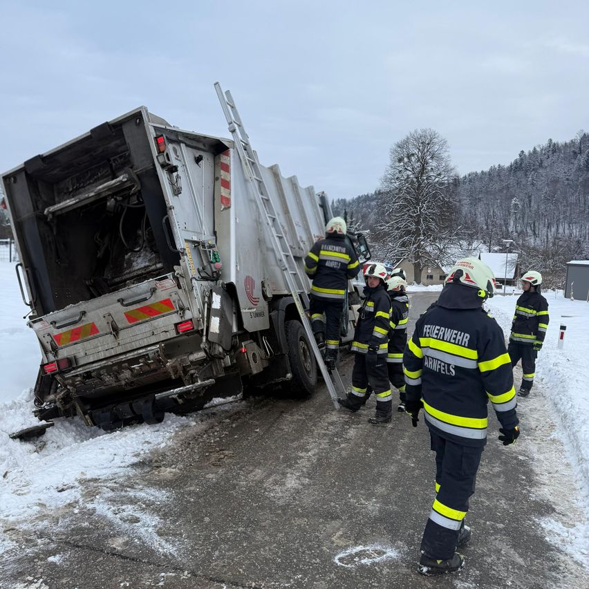 Feuerwehrleute kümmern sich um einen umgestürzten Lastwagen auf einer verschneiten Straße, wobei eine Person eine Leiter erklimmt und andere beobachten.