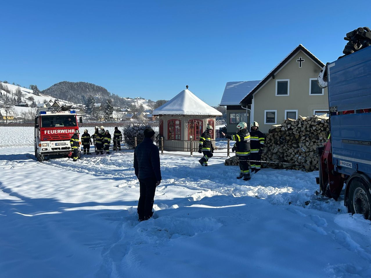 Feuerwehrleute in Schutzkleidung versammeln sich in einem verschneiten Bereich, nahe einem Feuerwehrwagen, einer kleinen Kapelle und einem Haus mit einem Kreuz auf dem Dach.