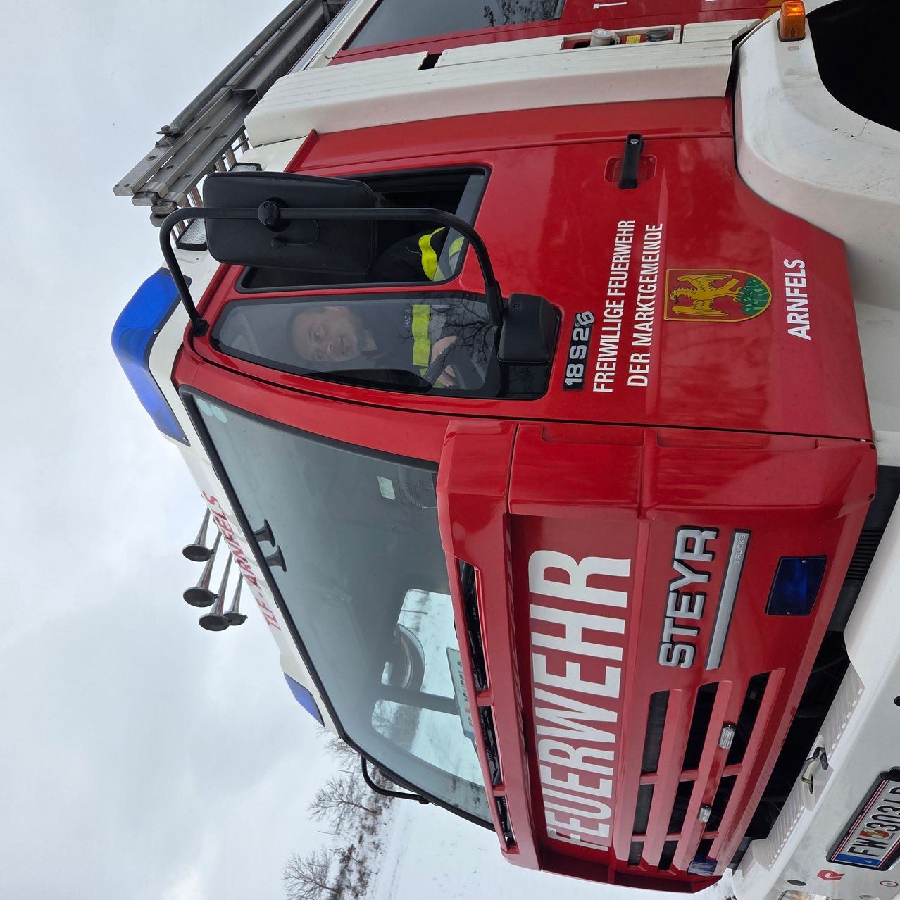 Ein Feuerwehrmann sitzt in einem roten Feuerwehrauto mit dem Wort 'Feuerwehr' darauf. Das Auto steht auf schneebedecktem Boden bei klarem Himmel.