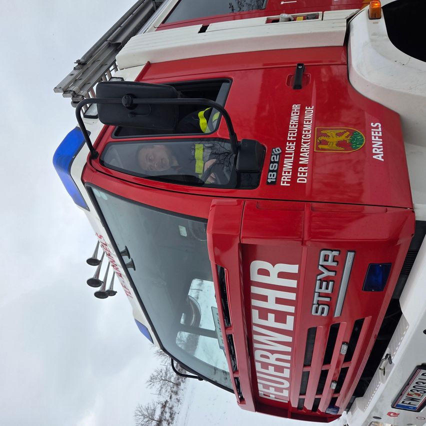 Ein Feuerwehrmann sitzt in einem roten Feuerwehrauto mit dem Wort 'Feuerwehr' darauf. Das Auto steht auf schneebedecktem Boden bei klarem Himmel.