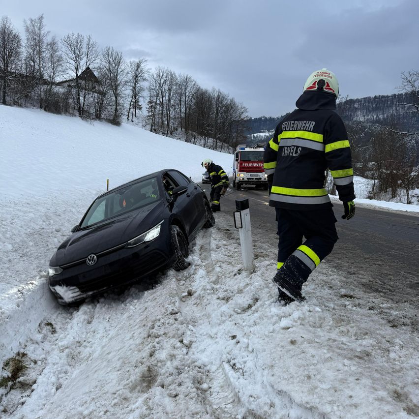 Ein schwarzer Volkswagen ist von der verschneiten Straße gerutscht und steckt im Schnee fest. Ein Feuerwehrmann nähert sich dem Auto mit einem weiteren Feuerwehrmann. Dahinter steht ein Feuerwehrwagen.