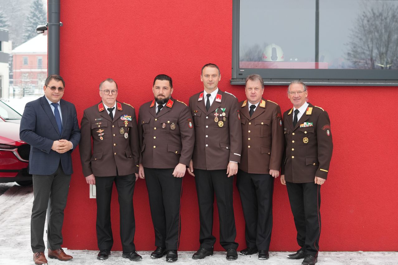 Fünf Männer in Militäruniformen stehen vor einer roten Wand, möglicherweise bei einer formellen Veranstaltung. Sie tragen Medaillen und Abzeichen an ihren Uniformen.