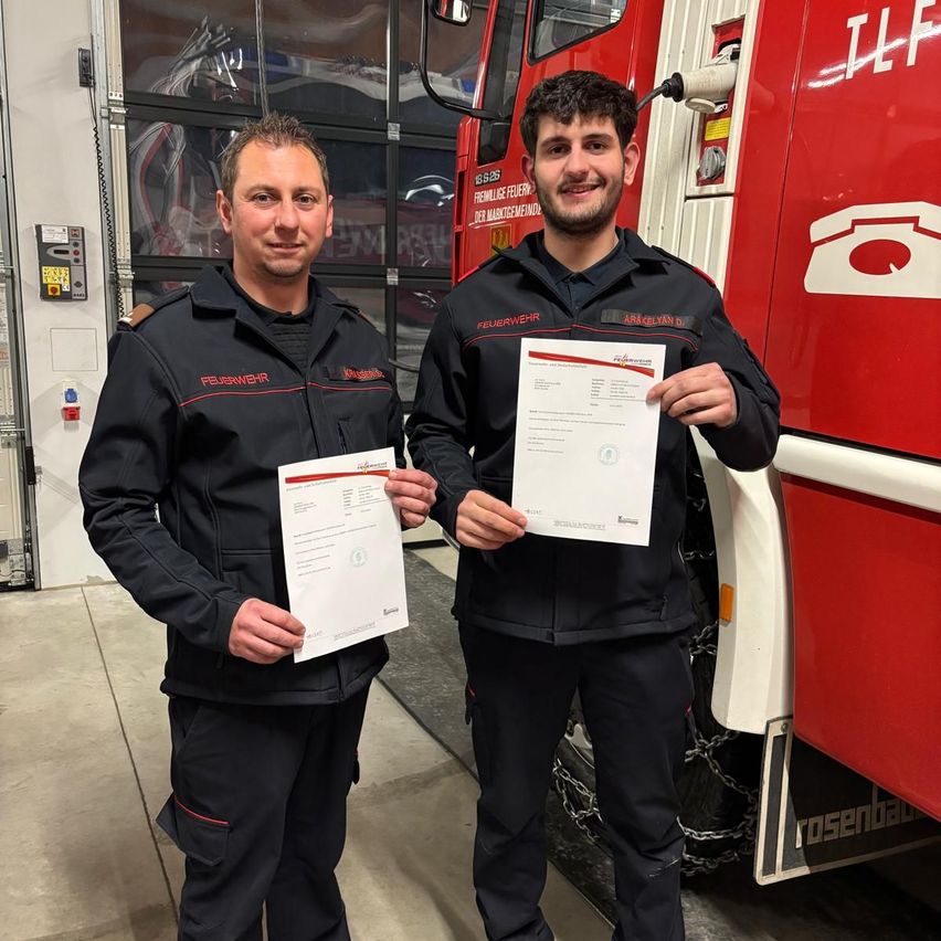 Zwei Feuerwehrleute posieren mit Urkunden vor einem Feuerwehrwagen in einer Garage.