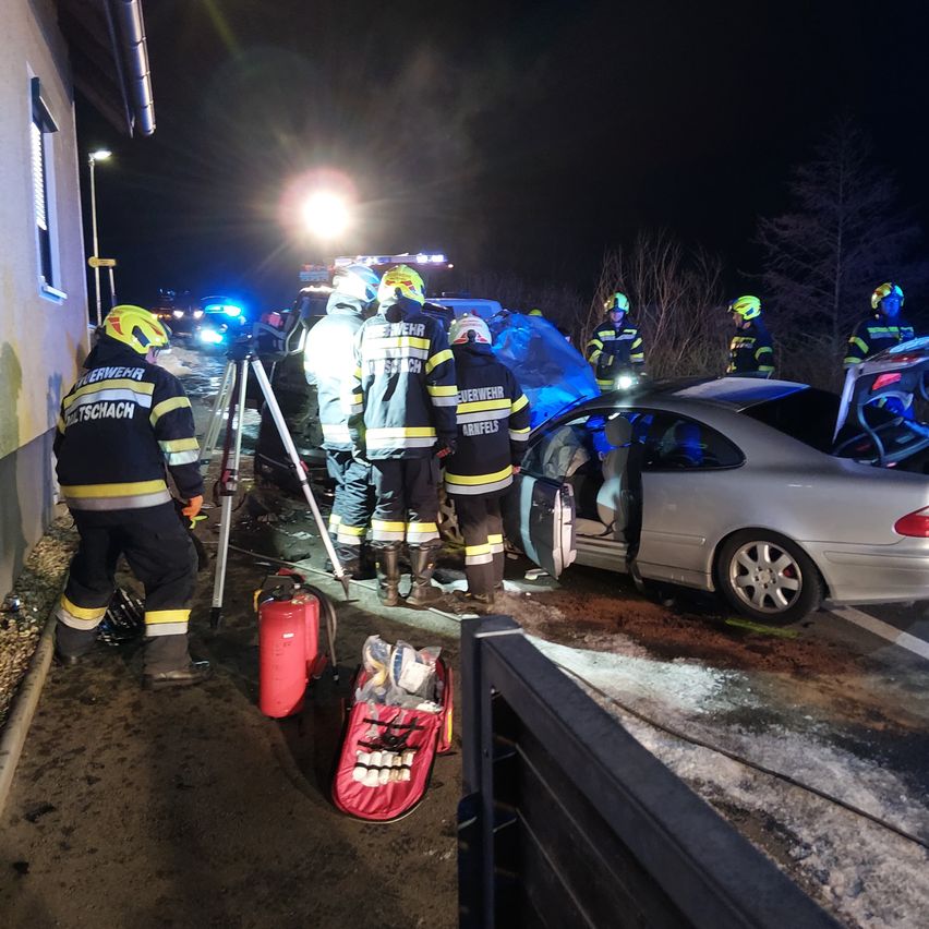 Feuerwehrleute in Schutzkleidung arbeiten bei einem Autounfall in der Nacht. Ein Auto ist beschädigt und seine Tür steht offen. Ausrüstung und Werkzeuge sind auf dem Boden verstreut.