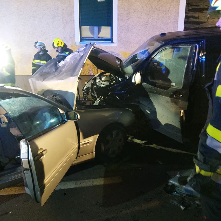 Ein Autounfall hat stattgefunden, mit einem Gebäude im Hintergrund. Rettungskräfte sind anwesend, und ein Auto hat einen beschädigten vorderen Teil und eine offene Tür.