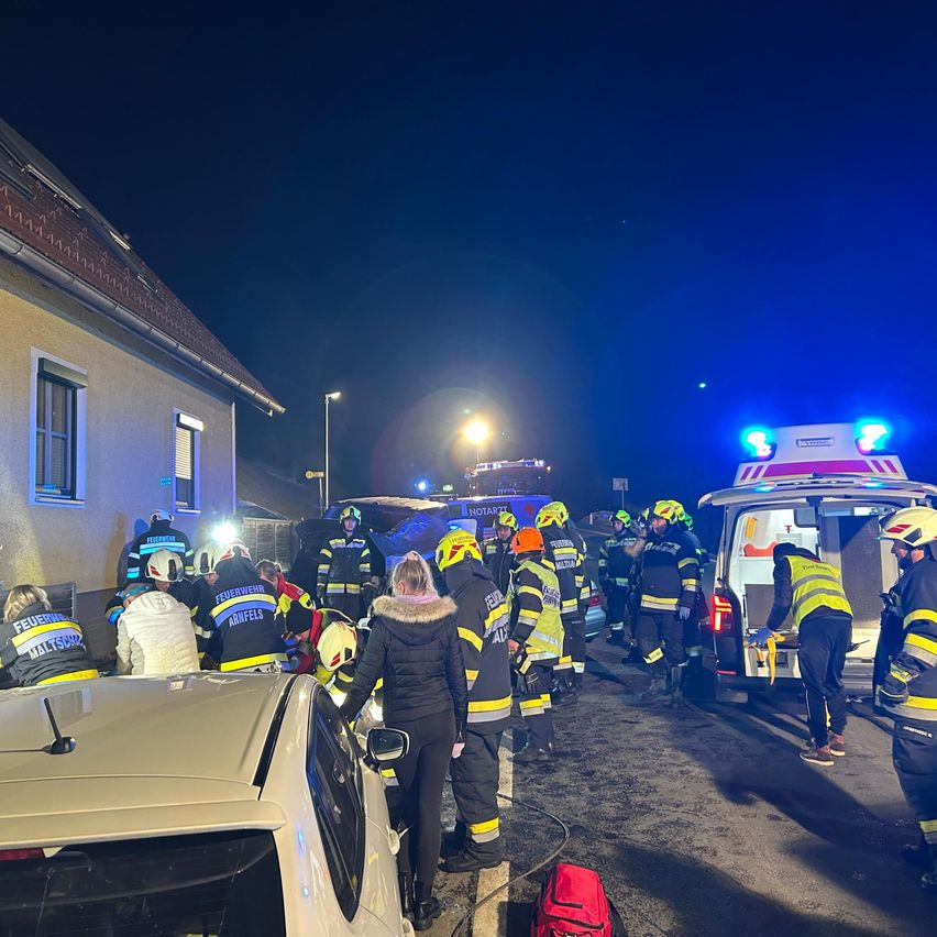 Einsatzkräfte versammeln sich an einem Unfallort bei Nacht. Ein Auto ist am Straßenrand geparkt, und ein Krankenwagen ist geöffnet. Feuerwehrleute versorgen die Verletzten.