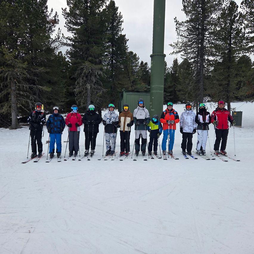 Eine Gruppe von Skifahrern und Snowboardern steht in einer Reihe auf einem verschneiten Hang, wobei im Hintergrund ein Skilift zu sehen ist.