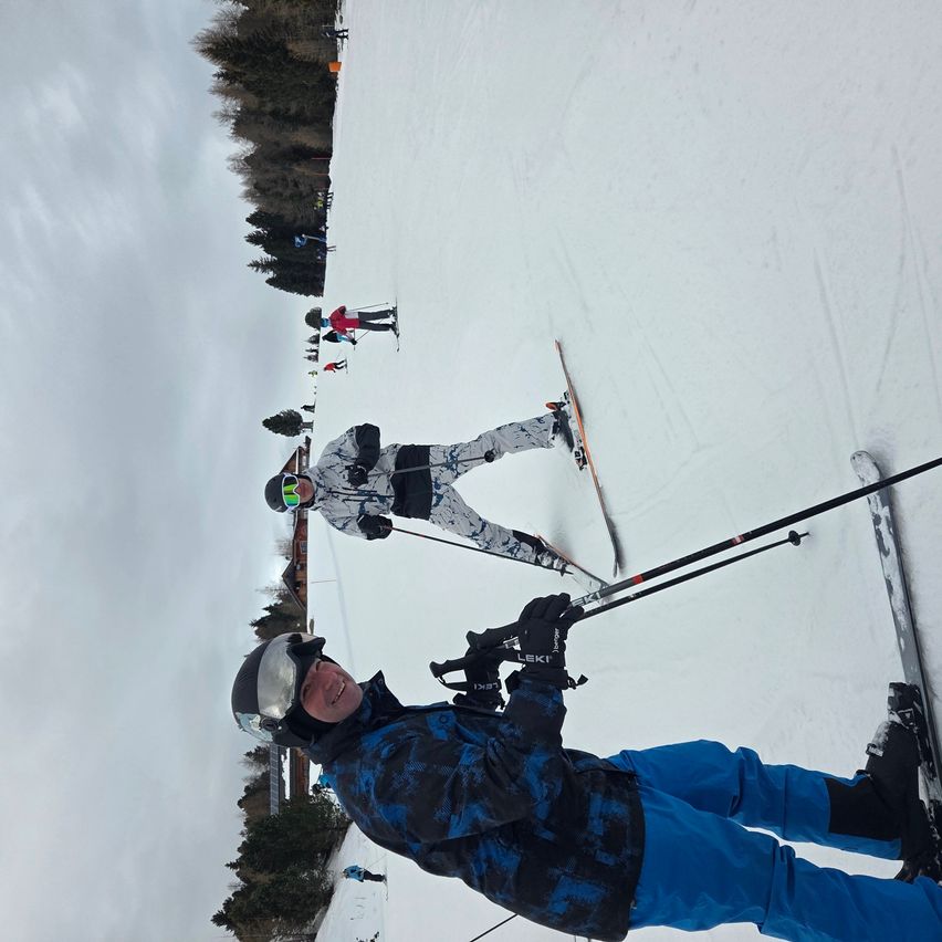 Ein Skifahrer auf einem verschneiten Hang, der lächelt und Skistöcke hält. Mehrere andere Skifahrer sind im Hintergrund zu sehen. Bäume und ein bewölkter Himmel sind sichtbar.