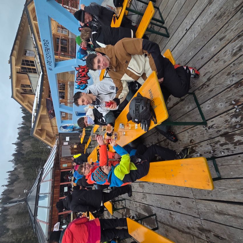 Eine Gruppe von Menschen sitzt an einem gelben Tisch in einem Skigebiet und trägt Winterkleidung. Einige trinken. Hinter ihnen steht ein Gebäude mit einer blauen Wand.
