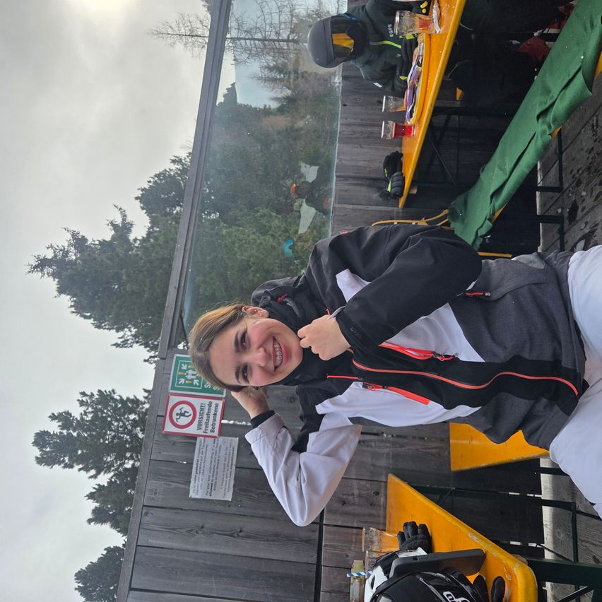 Eine Frau lächelt und posiert für ein Foto im Freien. Hinter ihr ist eine Holzstruktur mit einem Tisch und Stühlen zu sehen. Bäume und ein bewölkter Himmel im Hintergrund.