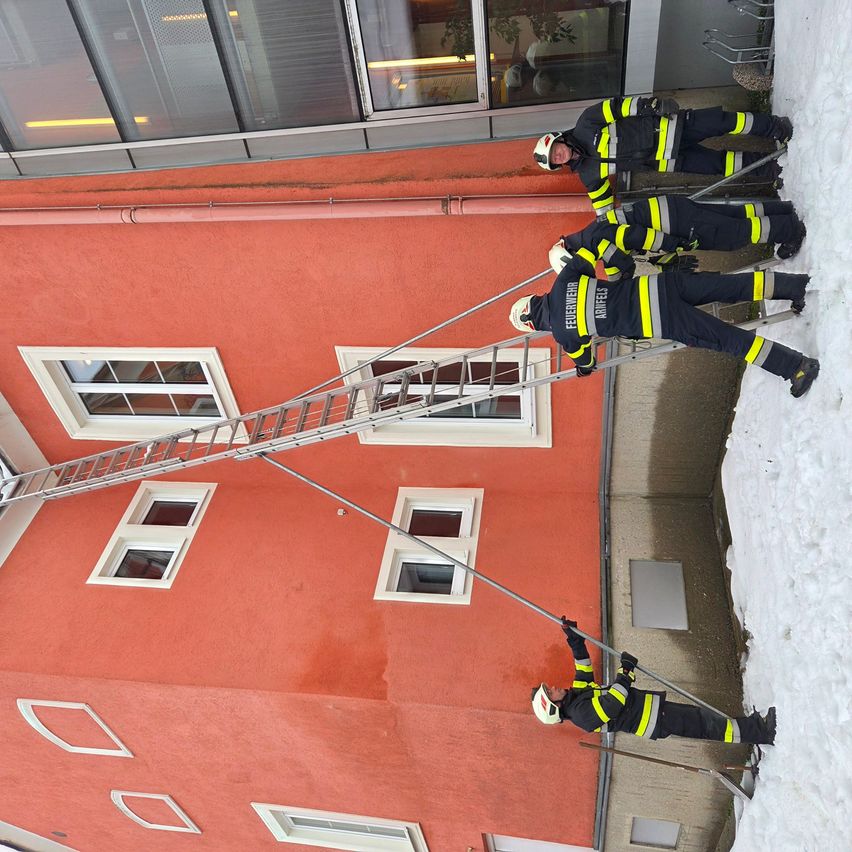Vier Feuerwehrleute in voller Montur klettern eine Leiter an einem roten Gebäude. Schnee liegt auf dem Boden und dem Dach.