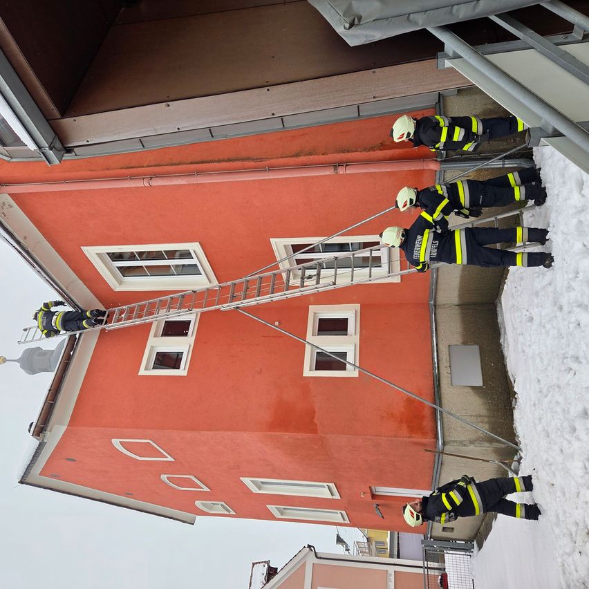 Eine Gruppe Feuerwehrleute in voller Ausrüstung klettert eine Leiter vor einem orangefarbenen Gebäude mit mehreren Fenstern hoch, an einem verschneiten Tag.