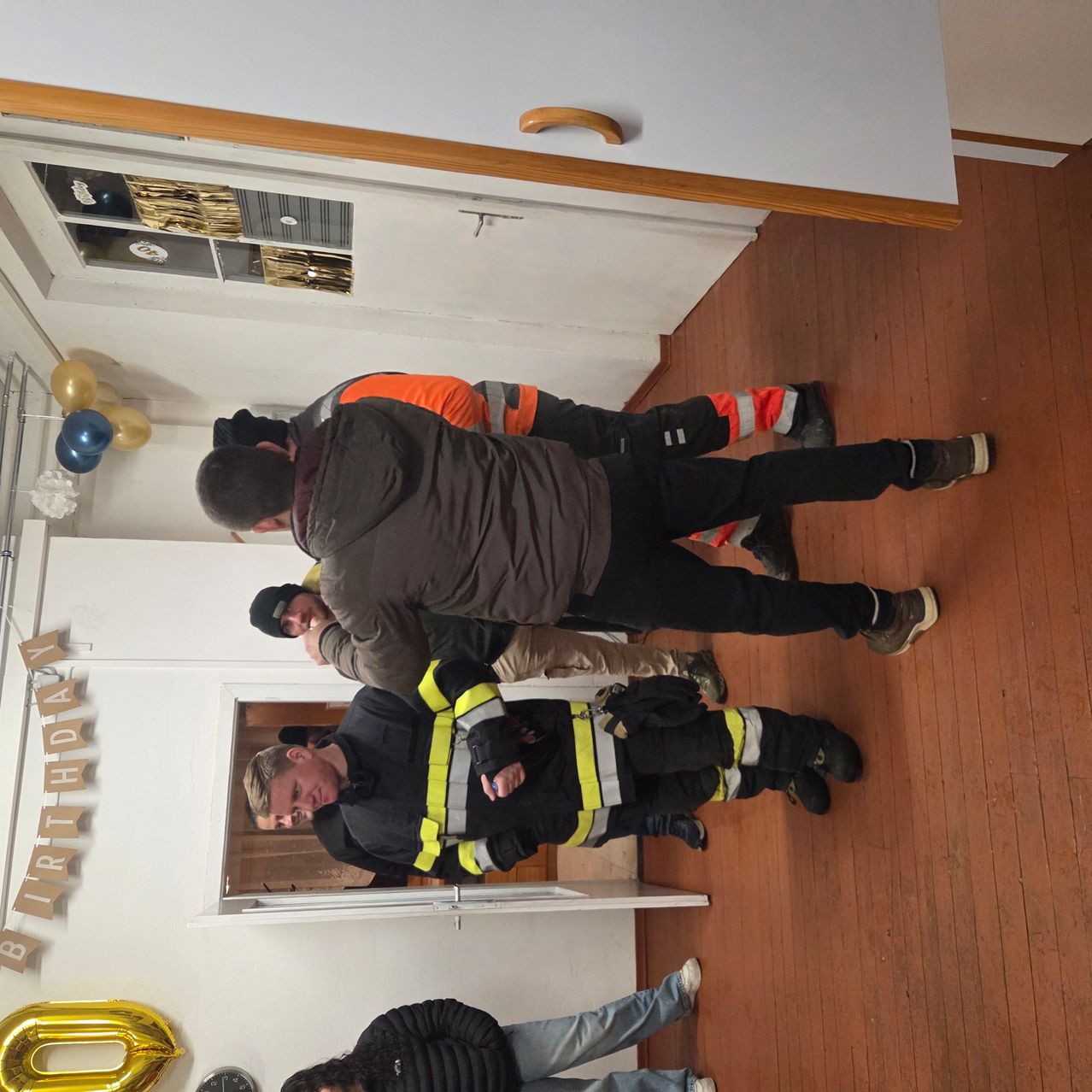 Eine Gruppe von Feuerwehrleuten steht in einem Raum und feiert einen Geburtstag. Im Hintergrund befinden sich Luftballons und eine Bannerfahne.