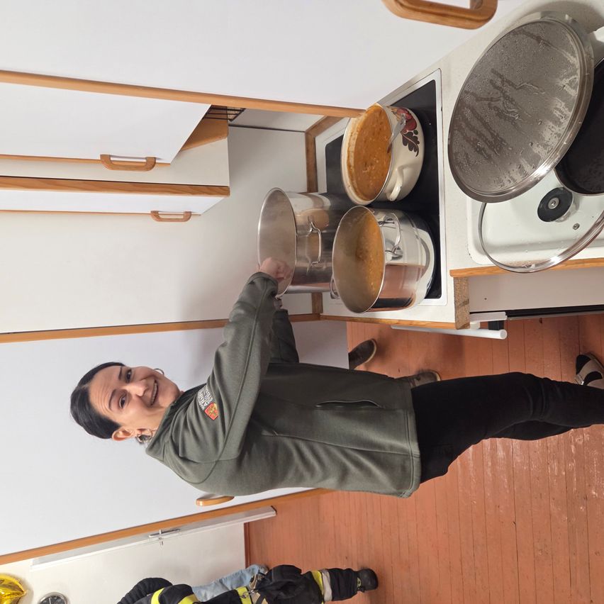 Eine Frau in einer Jacke lächelt, während sie einen Topf in einer Küche hebt. Sie steht vor einem Herd mit mehreren Töpfen, darunter einer mit einem Löffel.