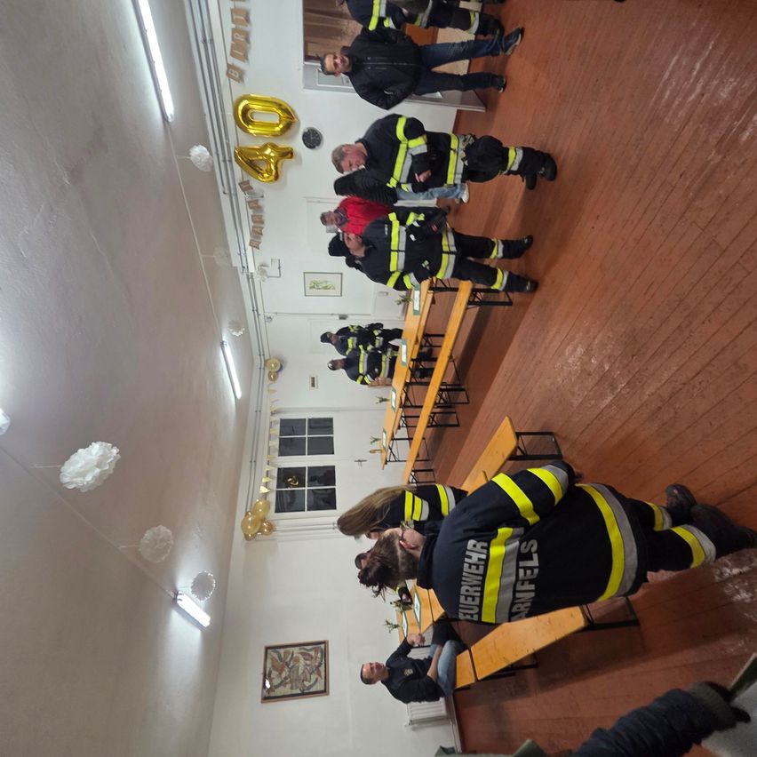 Eine Gruppe von Feuerwehrleuten in Uniform versammelt sich in einem Raum mit Holzböden und weißen Wänden. Es gibt Luftballons und Dekorationen. Ein Tisch mit Stühlen ist aufgebaut, und eine Person steht davor.