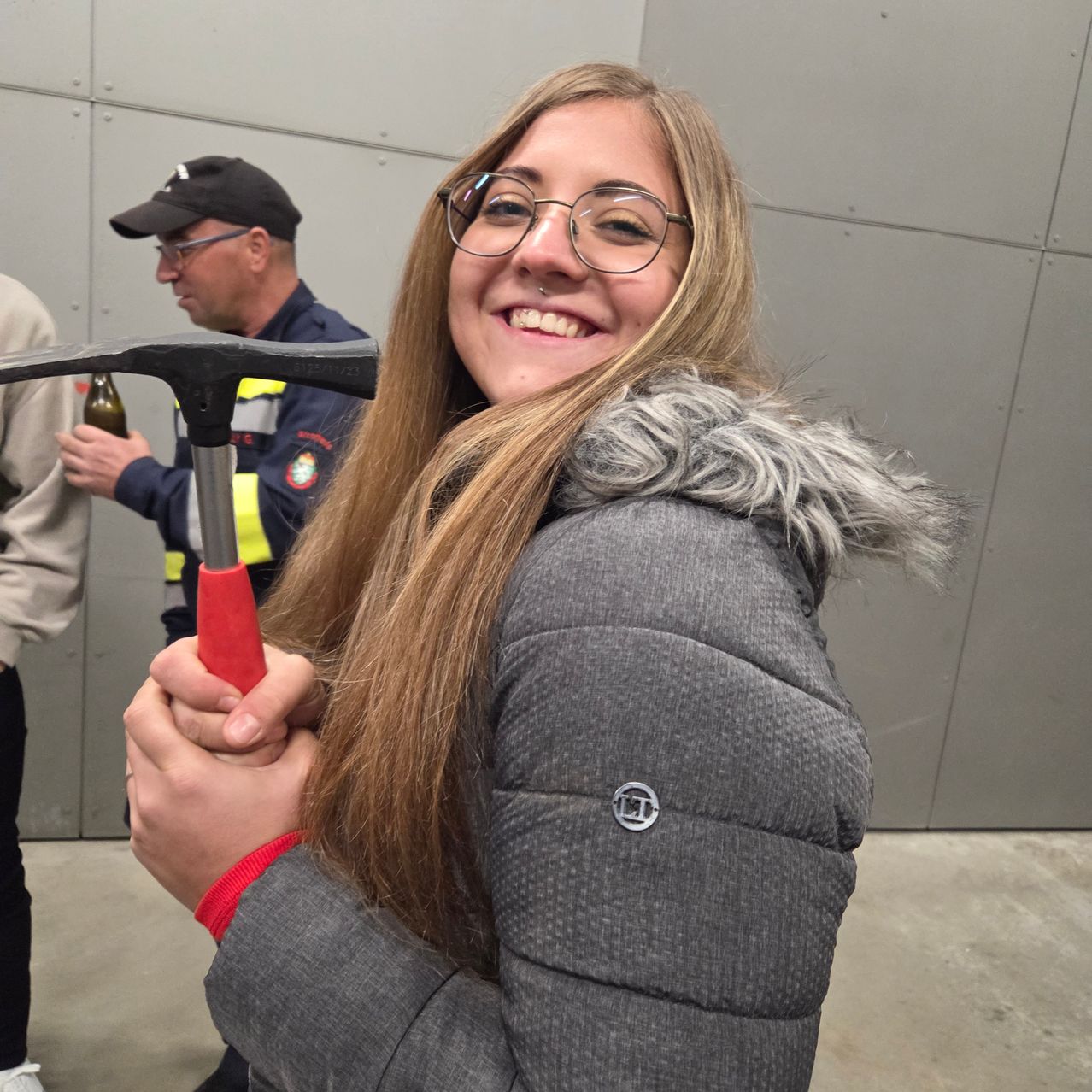 Eine Frau mit Brille und grauer Jacke lächelt und hält einen roten Hammer. Hinter ihr steht ein Mann in blauer Uniform mit gelber Weste, der eine Flasche hält. Eine dritte Person steht links.