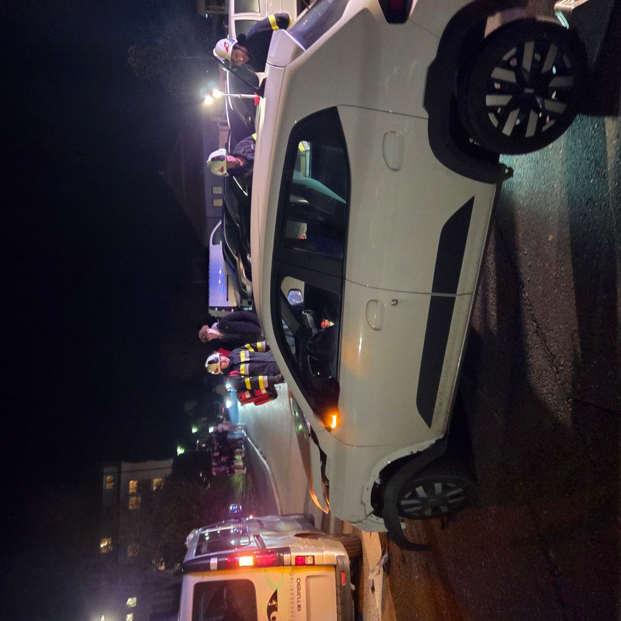 In der Nacht retten Feuerwehrleute eine Person aus einem weißen Auto mit offener Tür auf einer Straße.