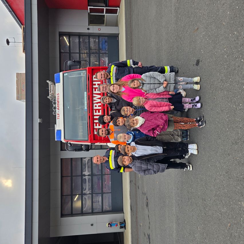 Eine Gruppe von Kindern und Erwachsenen posiert vor einem roten Feuerwehrauto. Das Wort 'Feuerwehr' steht auf dem Auto. Sie stehen auf einem gepflasterten Weg, und das Gebäude hinter ihnen hat Glasfenster.