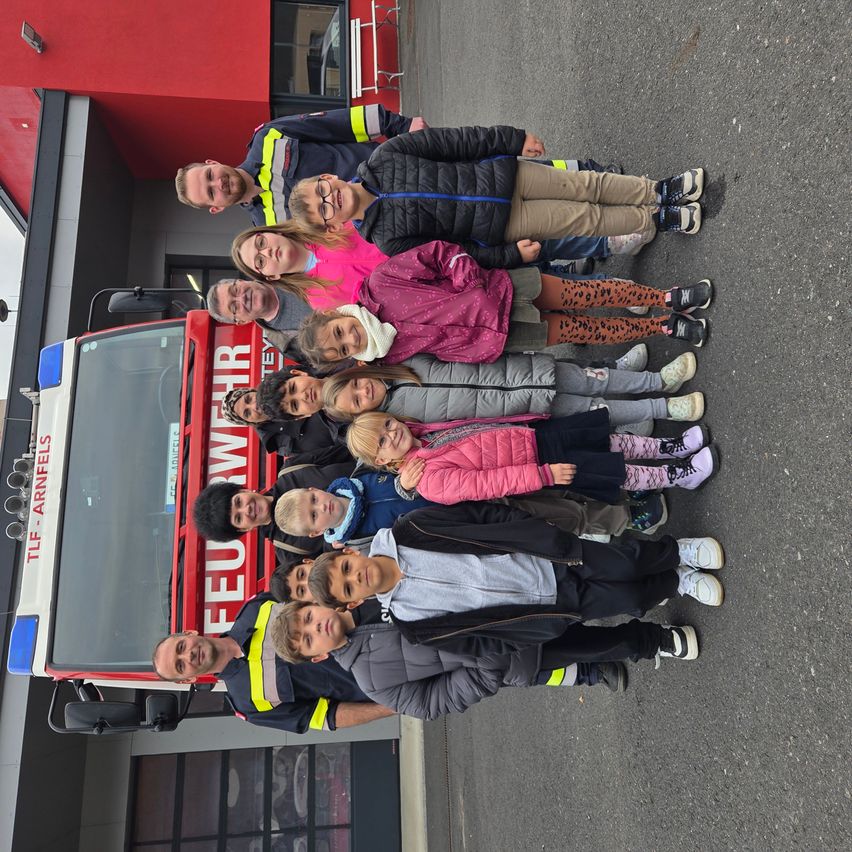 Gruppe von Kindern und Erwachsenen posiert vor einem roten Feuerwehrfahrzeug mit der Aufschrift TLF-Arnfels. Sie stehen auf einer gepflasterten Straße.