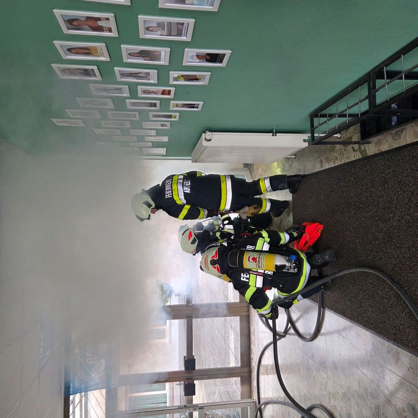 Feuerwehrleute trainieren in einem rauchgefüllten Flur mit Fotos an der Wand und einem Heizkörper darunter.