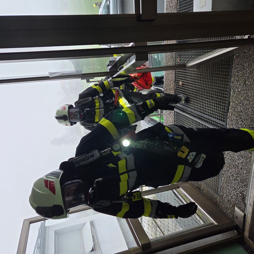 Drei Feuerwehrleute klettern eine Metalltreppe mit einem nebligen Hintergrund hoch. Sie tragen Helme und Schutzausrüstung. Die Person vorne hält eine Taschenlampe.