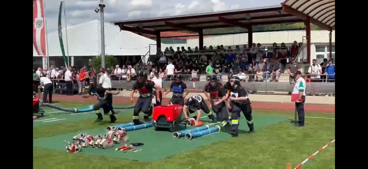 Several individuals dressed in protective gear and helmets are performing an activity on a field, possibly a firefighting simulation. Spectators are watching from a covered area.