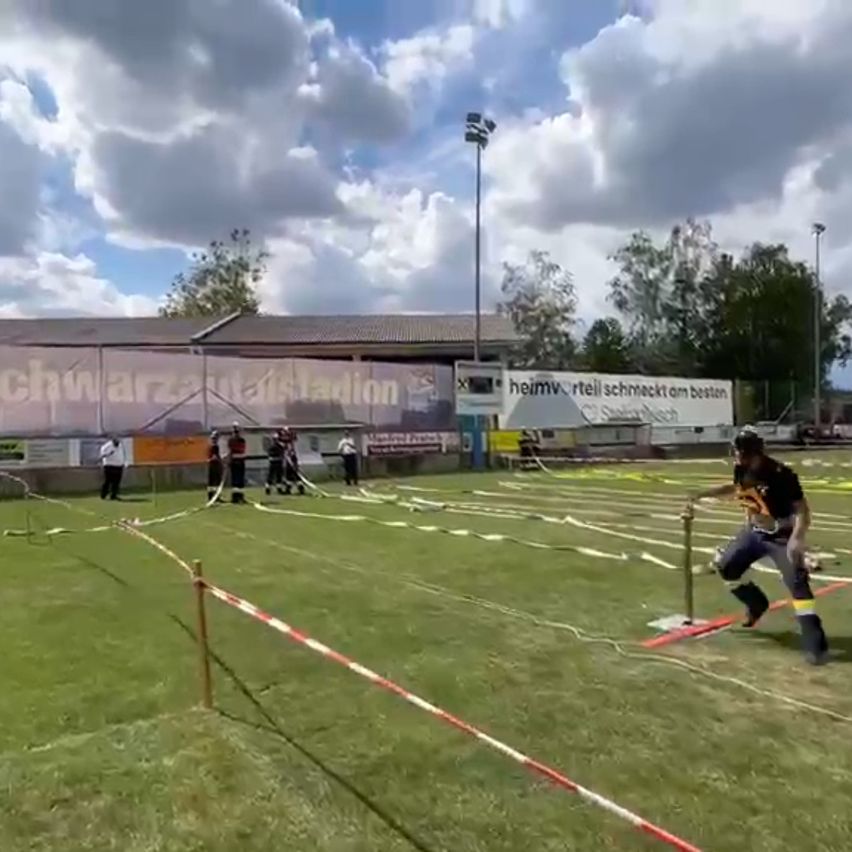 A man runs on a grassy field with hurdles and ropes. Spectators watch from behind the barriers. The sky is cloudy.