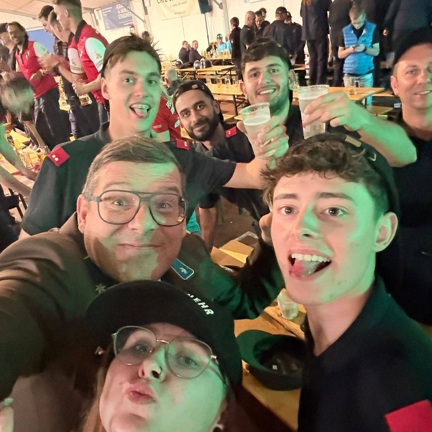 A group of young men in green uniforms with red patches are smiling and posing for a selfie at an outdoor event, holding drinks. Behind them, more people are standing and sitting around tables.